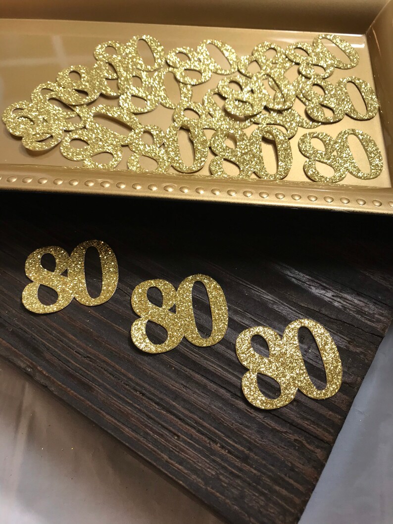 May include: Gold glitter confetti in the shape of the number 80. There are several pieces of confetti in a gold tray and three pieces of confetti on a dark brown wooden surface.