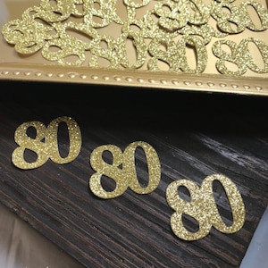 May include: Gold glitter number 80 cutouts. There are several 80s in a gold tray and three 80s on a dark wood surface.