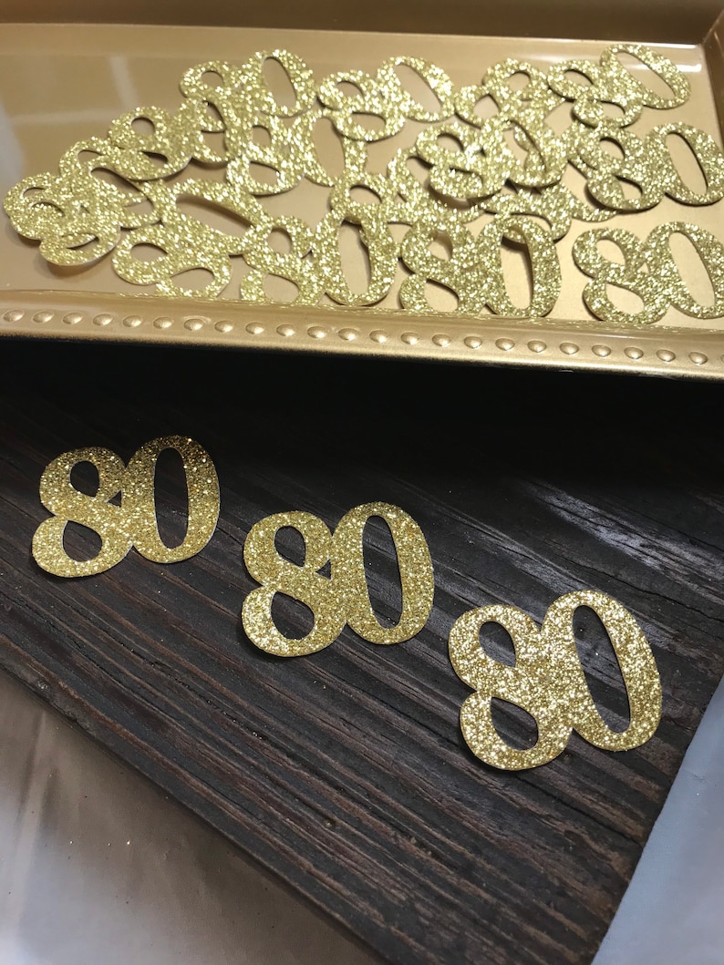 May include: Gold glitter number 80 cutouts. There are several 80s in a gold tray and three 80s on a dark wood surface.