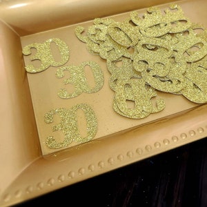 May include: A gold tray with a decorative border holds a pile of gold glitter paper cutouts of the number 30.