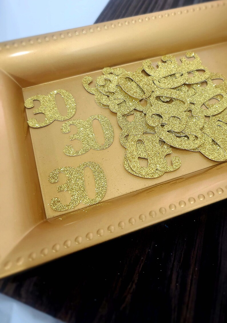 May include: A gold tray with a decorative border holds a pile of gold glitter paper cutouts of the number 30.