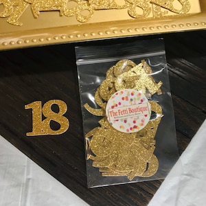 Puede incluir: Confetti de purpurina dorada en una bolsa de pl&aacute;stico transparente con el texto "The Fetti Boutique" y un n&uacute;mero 18 de purpurina dorada sobre una superficie de madera.