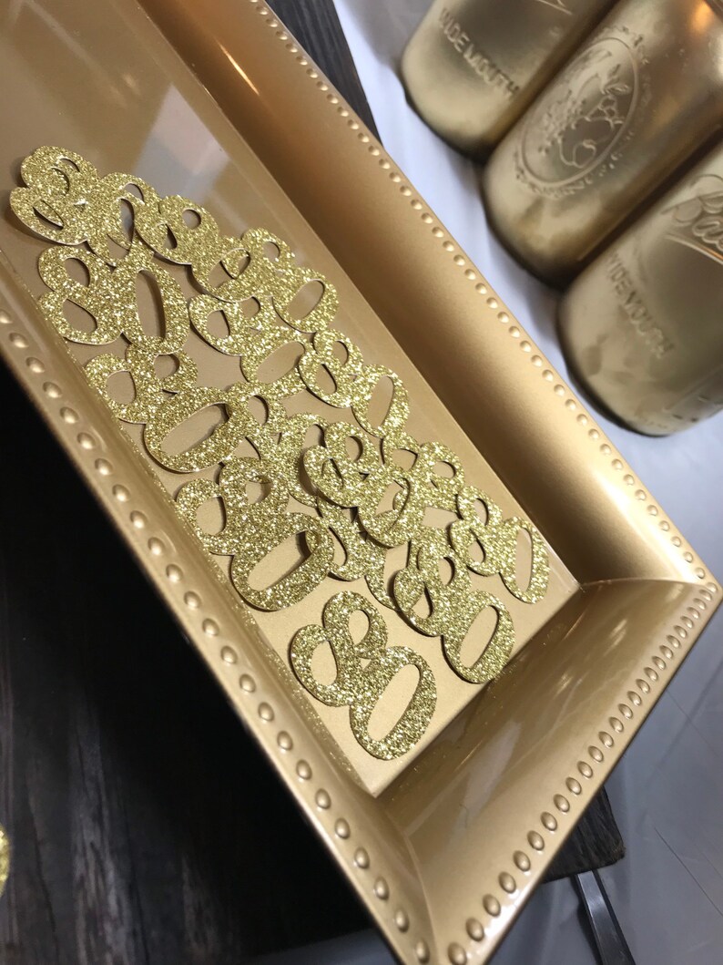 May include: A gold tray with a beaded border holds a scattering of gold glitter paper cutouts shaped like the number eight.