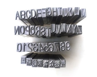 Pode incluir: Um conjunto de carimbos de metal para letras e números, incluindo o alfabeto, os números de 0 a 9 e vários caracteres chineses. Os carimbos estão dispostos em três filas sobre uma superfície branca.