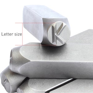 May include: Close-up of metal letter stamps. The stamp in focus displays the letter "K" within a circular indentation. The text "Letter size" is visible. The stamps are stacked, showing their rectangular bodies and rounded edges.