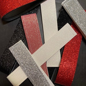 May include: Assortment of glitter fabric strips in red, silver, white, black, and pink. The strips are arranged in a criss-cross pattern on a black background.