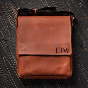 May include: A brown leather messenger bag with a flap closure and a personalized monogram that reads "EIW".