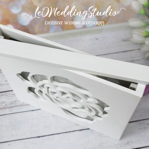 May include: A rectangular white wooden box with a hinged lid featuring a laser-cut rose design on the front panel. The box has a smooth matte finish and is positioned at an angle to show the top lid and the intricate floral cutout detail.