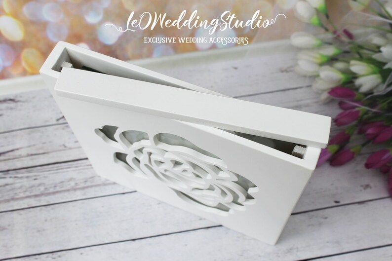 May include: A rectangular white wooden box with a hinged lid featuring a laser-cut rose design on the front panel. The box has a smooth matte finish and is positioned at an angle to show the top lid and the intricate floral cutout detail.