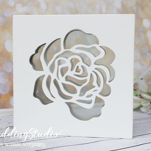 May include: A square white box with a cutout of a rose flower on the front face. The text "leWeddingStudio" and "EXCLUSIVE WEDDING ACCESSORIES" is printed in white on the lower left corner of the box, which features a layered design.