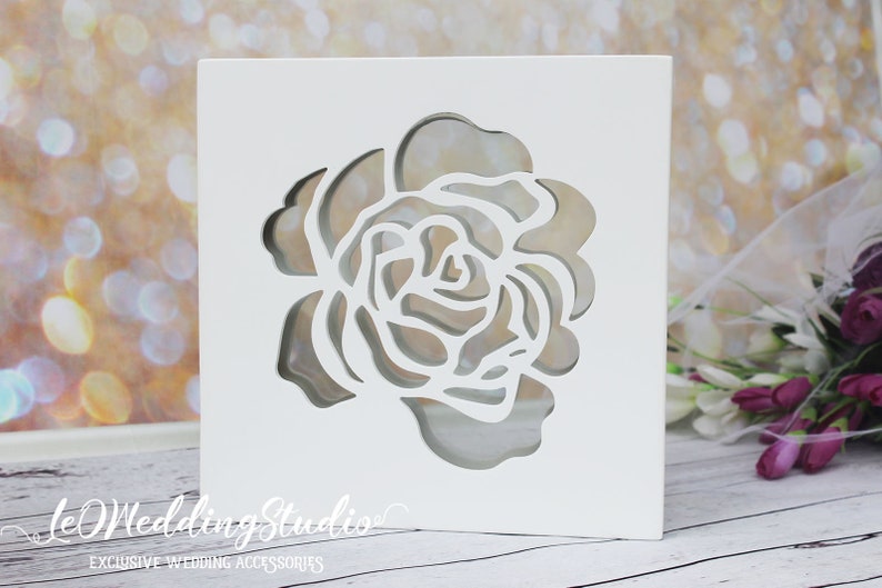 May include: A square white box with a cutout of a rose flower on the front face. The text "leWeddingStudio" and "EXCLUSIVE WEDDING ACCESSORIES" is printed in white on the lower left corner of the box, which features a layered design.