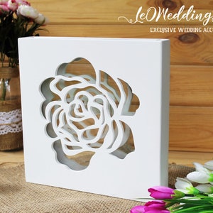 May include: A white square wooden box featuring a large, intricate laser-cut rose design in the center. The text "leWeddingStudio EXCLUSIVE WEDDING ACCESSORIES" is printed in white in the upper right corner of the image.