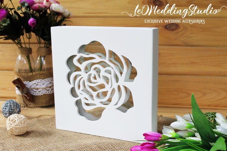 May include: A white square wooden box featuring a large, intricate laser-cut rose design in the center. The text "leWeddingStudio EXCLUSIVE WEDDING ACCESSORIES" is printed in white in the upper right corner of the image.