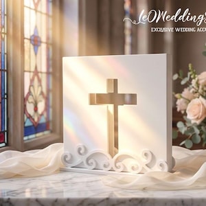 May include: A white square plaque featuring a central cutout cross and decorative scrollwork at the base. The plaque is positioned on a marble surface with draped fabric. The text "leWeddingStudio EXCLUSIVE WEDDING ACCESSORIES" is visible.