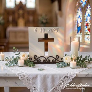 May include: A white square wooden sign featuring a cutout cross in the center and the date 6 12 2025 in raised numbers. The base is decorated with dark brown scrollwork, and the sign is surrounded by white roses and eucalyptus leaves.