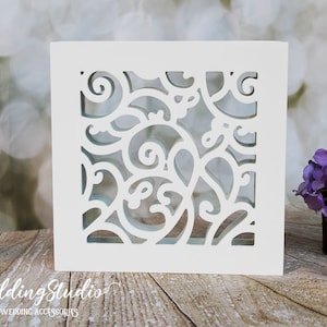 May include: White square card with a decorative cut-out design of swirling vines. The card is on a wooden surface with a purple flower arrangement and a ball of twine.