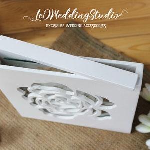 May include: A white wooden box with a sliding lid featuring a laser-cut rose design on the front panel. The text "leOWeddingStudio" and "EXCLUSIVE WEDDING ACCESSORIES" is printed in white above the box on the wooden surface.