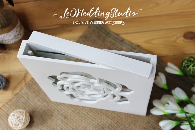 May include: A white wooden box with a sliding lid featuring a laser-cut rose design on the front panel. The text "leOWeddingStudio" and "EXCLUSIVE WEDDING ACCESSORIES" is printed in white above the box on the wooden surface.