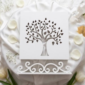 Peut inclure: Une plaque carrée en bois blanc présentant une silhouette d'arbre découpée au laser avec des feuilles et des branches brun foncé. La plaque repose sur un support blanc décoratif aux motifs de volutes complexes, centré sur un tissu blanc.