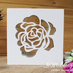 May include: A square white wooden box featuring a laser-cut rose design on the front panel. The cutout reveals the interior space through the floral pattern. The text "leWeddingStudio" and "EXCLUSIVE WEDDING ACCESSORIES" is printed in white.