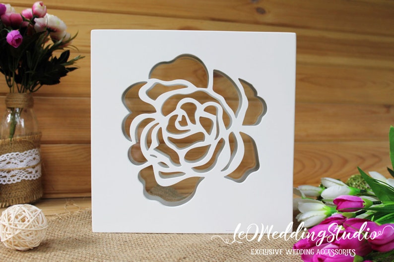 May include: A square white wooden box featuring a laser-cut rose design on the front panel. The cutout reveals the interior space through the floral pattern. The text "leWeddingStudio" and "EXCLUSIVE WEDDING ACCESSORIES" is printed in white.