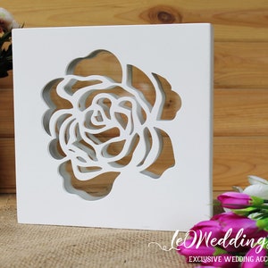 May include: A square white wooden block featuring a laser-cut rose design in the center. The cutout creates a hollow floral silhouette through the material. The text LeWeddingStudio EXCLUSIVE WEDDING ACCESSORIES is printed in white at the bottom right.