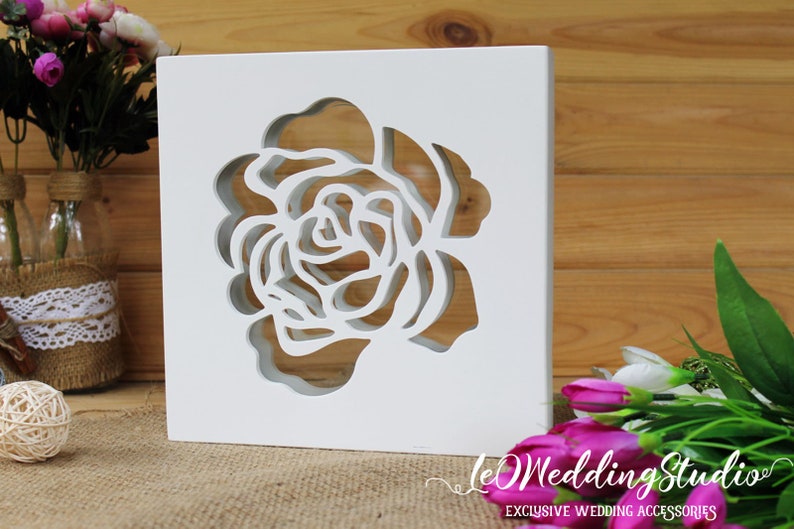 May include: A square white wooden block featuring a laser-cut rose design in the center. The cutout creates a hollow floral silhouette through the material. The text LeWeddingStudio EXCLUSIVE WEDDING ACCESSORIES is printed in white at the bottom right.
