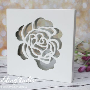 May include: A square white wooden box featuring a laser-cut rose design on the front panel. The cutout reveals a hollow interior space. The text "leWeddingStudio" and "EXCLUSIVE WEDDING ACCESSORIES" is printed in white at the bottom left corner.