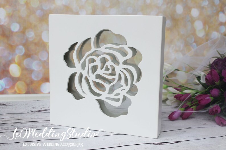 May include: A square white wooden box featuring a laser-cut rose design on the front panel. The cutout reveals a hollow interior space. The text "leWeddingStudio" and "EXCLUSIVE WEDDING ACCESSORIES" is printed in white at the bottom left corner.