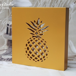 May include: A gold square wooden box with a cutout pineapple design. The pineapple is in the centre of the box and is a light brown colour.