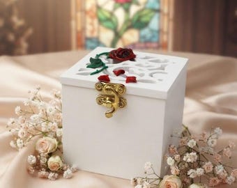May include: A square white wooden box featuring a gold-toned metal latch on the front. The lid has a cutout design decorated with a small red rose and green leaves. The text LeWeddingStudio EXCLUSIVE WEDDING ACCESSORIES is printed at the bottom.