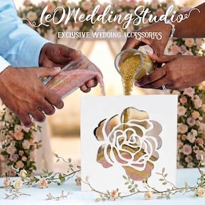 May include: A white square sand ceremony frame with a cutout rose design on the front. Two glass vessels are pouring pink and gold sand into the frame. The text "leOWeddingStudio EXCLUSIVE WEDDING ACCESSORIES" is visible at the top of the image.