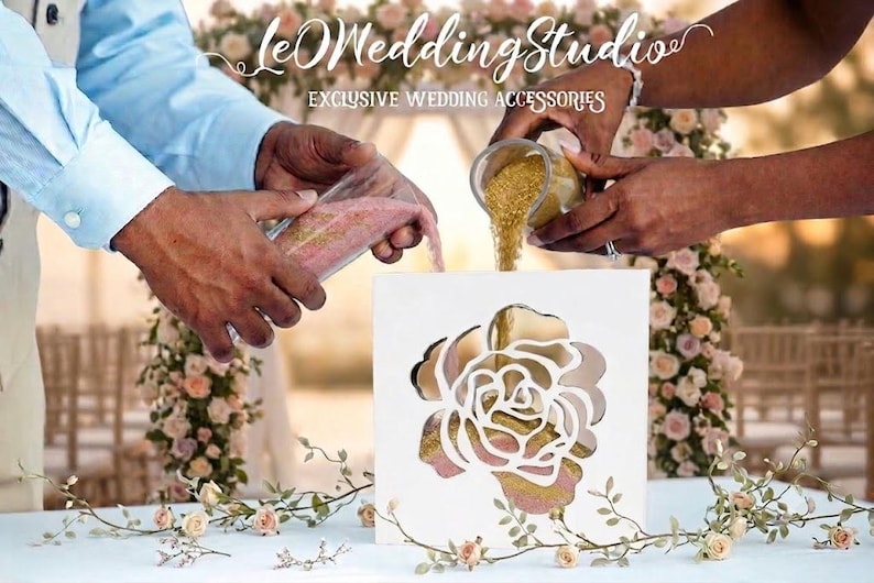 May include: A white square sand ceremony frame with a cutout rose design on the front. Two glass vessels are pouring pink and gold sand into the frame. The text "leOWeddingStudio EXCLUSIVE WEDDING ACCESSORIES" is visible at the top of the image.