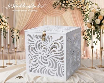 May include: A white square card box featuring intricate laser-cut swirling patterns on its sides and top. The box includes a gold-colored metal latch with a small padlock attached to the front, and a set of keys is placed on the table nearby.