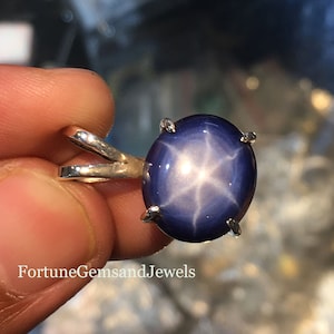 May include: A blue star sapphire ring with a silver band. The sapphire has a six-pointed star pattern in the center. The ring is held up by a hand. The text "Fortune Gems and Jewels" is at the bottom.