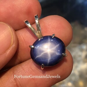 May include: A blue star sapphire pendant with a silver bail. The gemstone has a six-rayed star pattern. The pendant is held in a hand. The text "Fortune Gems and Jewels" is visible at the bottom.