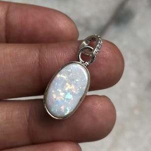 May include: An oval-shaped pendant featuring a white opal gemstone with iridescent flashes of color, set in a polished silver-toned metal frame. The pendant includes a small, circular silver-toned bail at the top for attachment to a chain or cord.