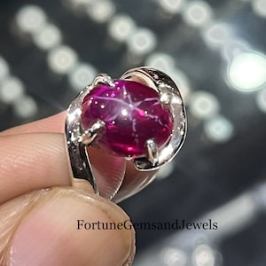 May include: A silver ring with a large, oval-cut, deep red gemstone exhibiting a star effect. The gemstone is held in a claw setting. The ring is shown against a blurred background.