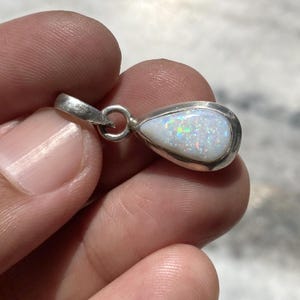 May include: A teardrop-shaped pendant featuring a white opal with iridescent flashes of color set in a smooth silver frame. The pendant includes a small circular silver bail at the top for attachment to a chain, showcasing a polished metal finish.