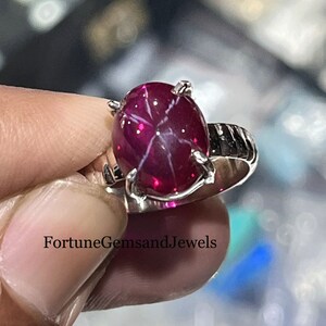 May include: A silver ring featuring a large, oval-shaped, star ruby gemstone. The ruby is a deep red color with a visible star effect. The ring has a simple, elegant design with four prongs holding the stone.