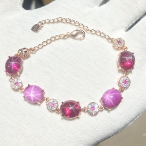 Può includere: Bracciale in oro rosa con pietre preziose di rubino ovali a forma di stella e pietre preziose chiare più piccole. Il bracciale ha una catena delicata e una chiusura. I rubini sono di colore rosso scuro e rosa.