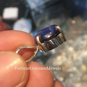 May include: A close-up of a pendant featuring a large, oval-shaped, deep blue gemstone with a star-like pattern. The gemstone is set in a silver-colored metal setting. The pendant is held by a person's fingers. The text "FortuneGemsandJewels" is visible.