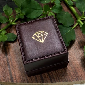 May include: A closed, rectangular jewelry box in a dark burgundy color. The box has a gold diamond-shaped emblem on the top. The box is sitting on a wooden surface with green leaves in the background.
