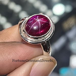 May include: A silver ring featuring a large, oval, deep red gemstone with a star-like pattern. The ring's band has a unique, swirling design. The text "Fortune Gems and Jewels" is visible on the ring.