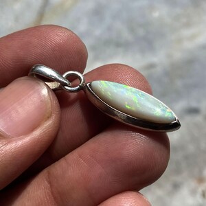 May include: A marquise-shaped pendant featuring a smooth, white opal gemstone with iridescent green and blue flashes, set in a polished silver-toned metal bezel with a circular bail at the top for attachment to a necklace chain.