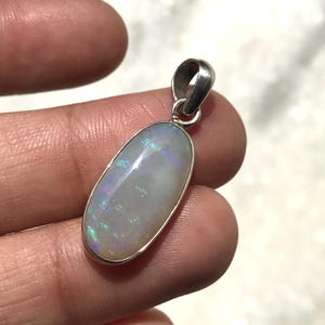 May include: An oval-shaped pendant featuring a smooth, milky white opal gemstone with iridescent flashes of blue and green light. The stone is set in a polished silver-toned metal bezel with a matching silver-toned metal bail at the top.
