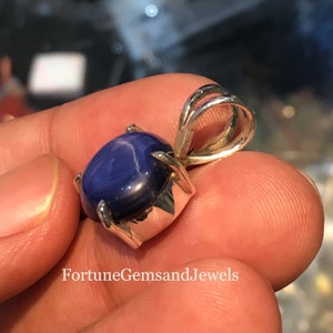 May include: A blue star sapphire pendant with a silver setting. The gemstone is oval-shaped and has a star-like effect. The pendant is held up by a silver bail. The text "FortuneGemsandJewels" is at the bottom of the image.