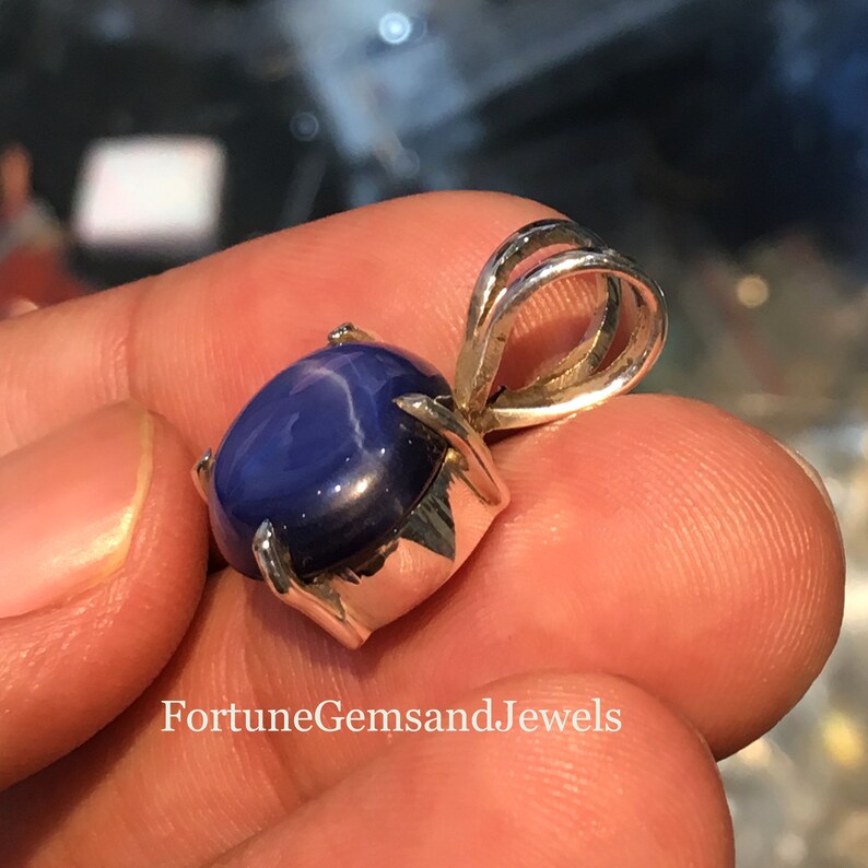 May include: A blue star sapphire pendant with a silver setting. The gemstone is oval-shaped and has a star-like effect. The pendant is held up by a silver bail. The text "FortuneGemsandJewels" is at the bottom of the image.