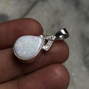 May include: A teardrop-shaped white opal pendant set in a silver-toned metal frame. The pendant features a bail accented with small, clear, round-cut stones. The opal displays iridescent flashes of red, green, and blue color throughout its surface.