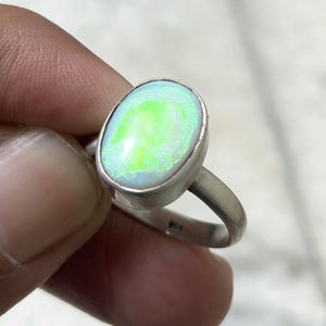 May include: A silver ring featuring an oval-shaped cabochon opal with a prominent neon green play-of-color. The stone is set in a smooth, polished silver bezel. The inner band of the ring is clearly stamped with the hallmark 925, indicating sterling silver.